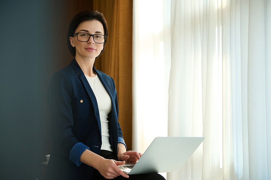 Business portrait of a beautiful self-confident young businesswoman, successful sales manager, investor, female investment advisor wearing eyeglasses, holding laptop and confidently looking at camera